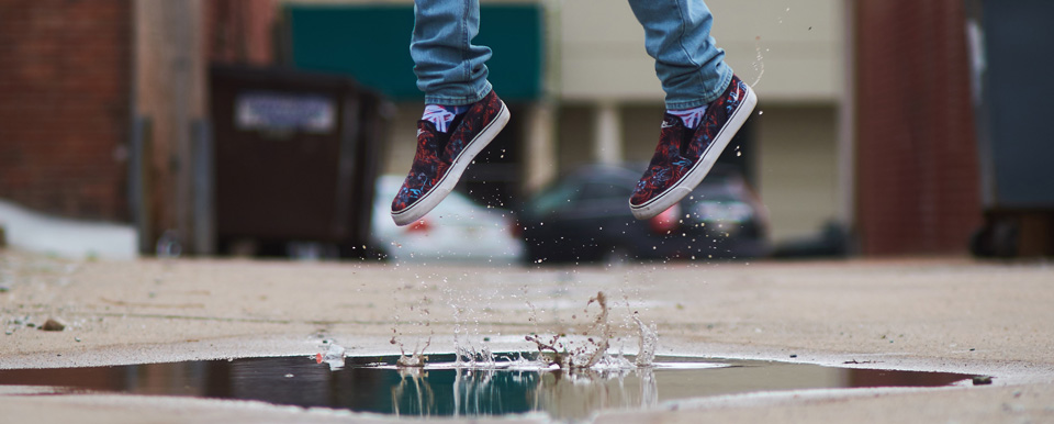 Man sieht Füße mit Schuhen, die aus einer Pfütze hochspringen.